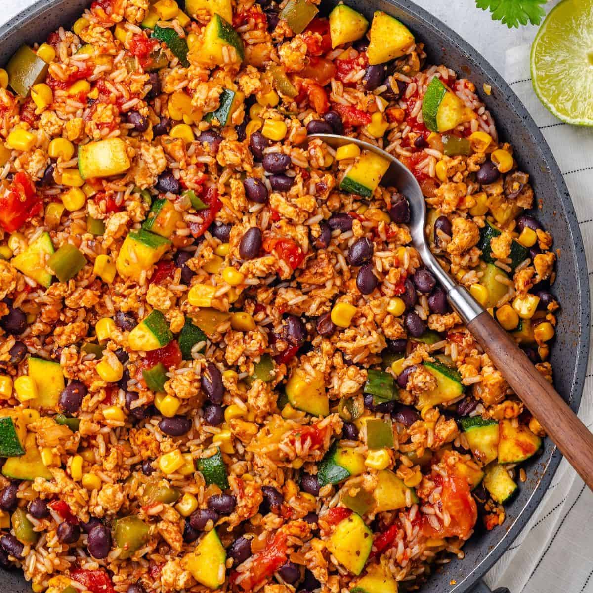 Close up of a spoon resting in a colorful skillet of ground meat with rice, beans and veggies.