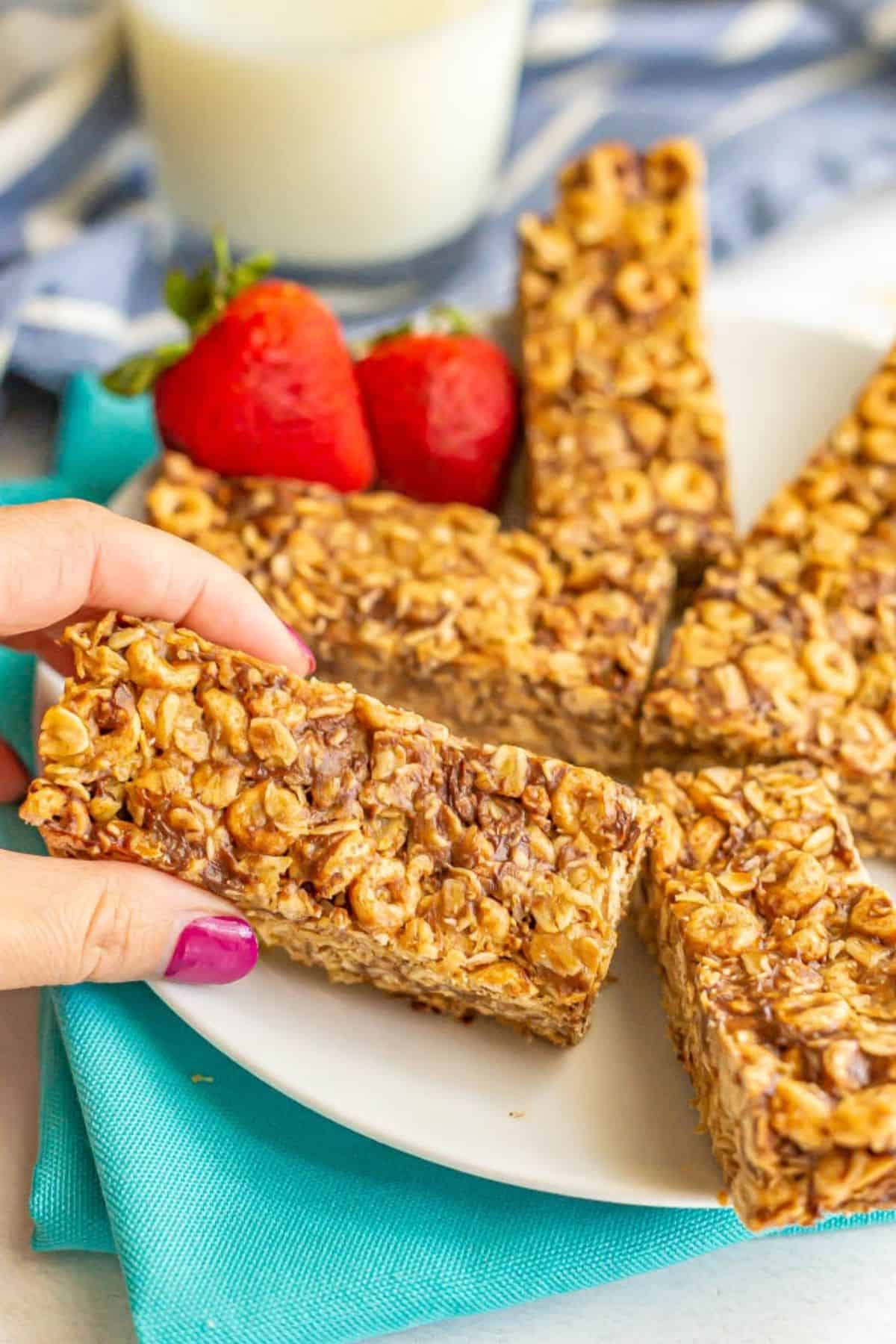 A hand picking up a no bake oat and cereal breakfast bar from a plate of bars with a glass of milk in the background.