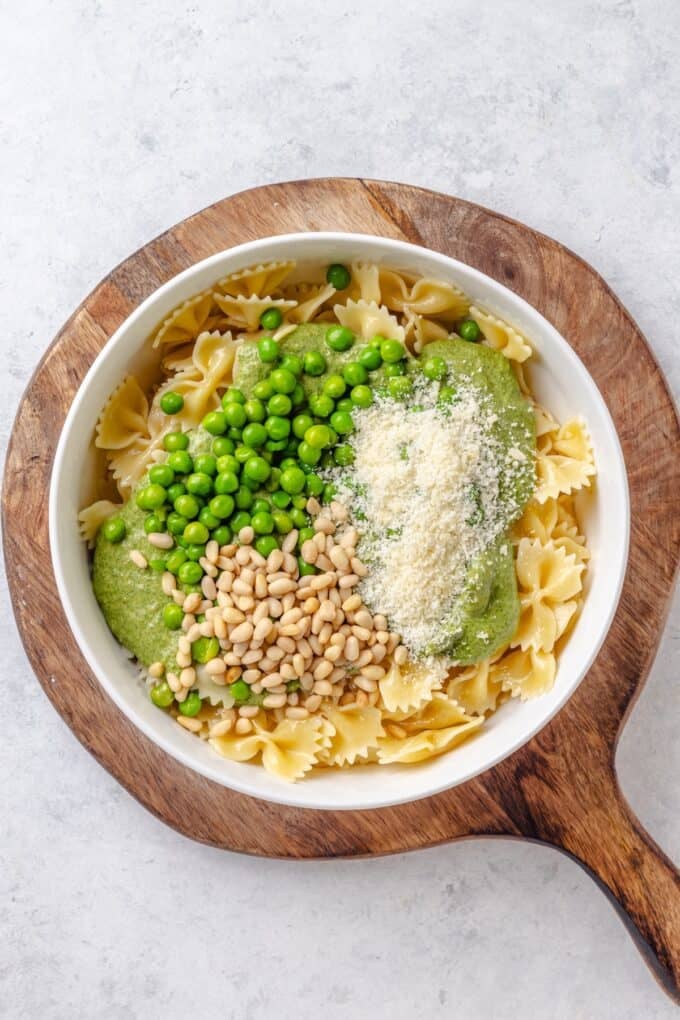 A large white bowl with cooked pasta topped with piles of a creamy pesto mixture, green peas, pine nuts and Parmesan cheese.
