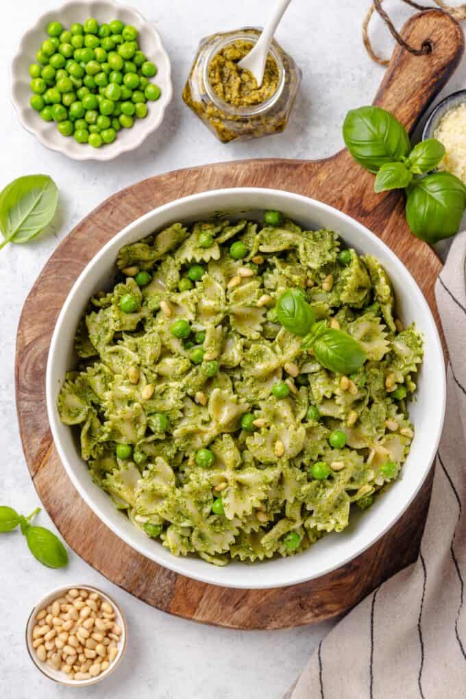 A large white serving bowl set on a cutting board filled with a pasta salad with pesto, peas, pine nuts and fresh basil on top and to the side.