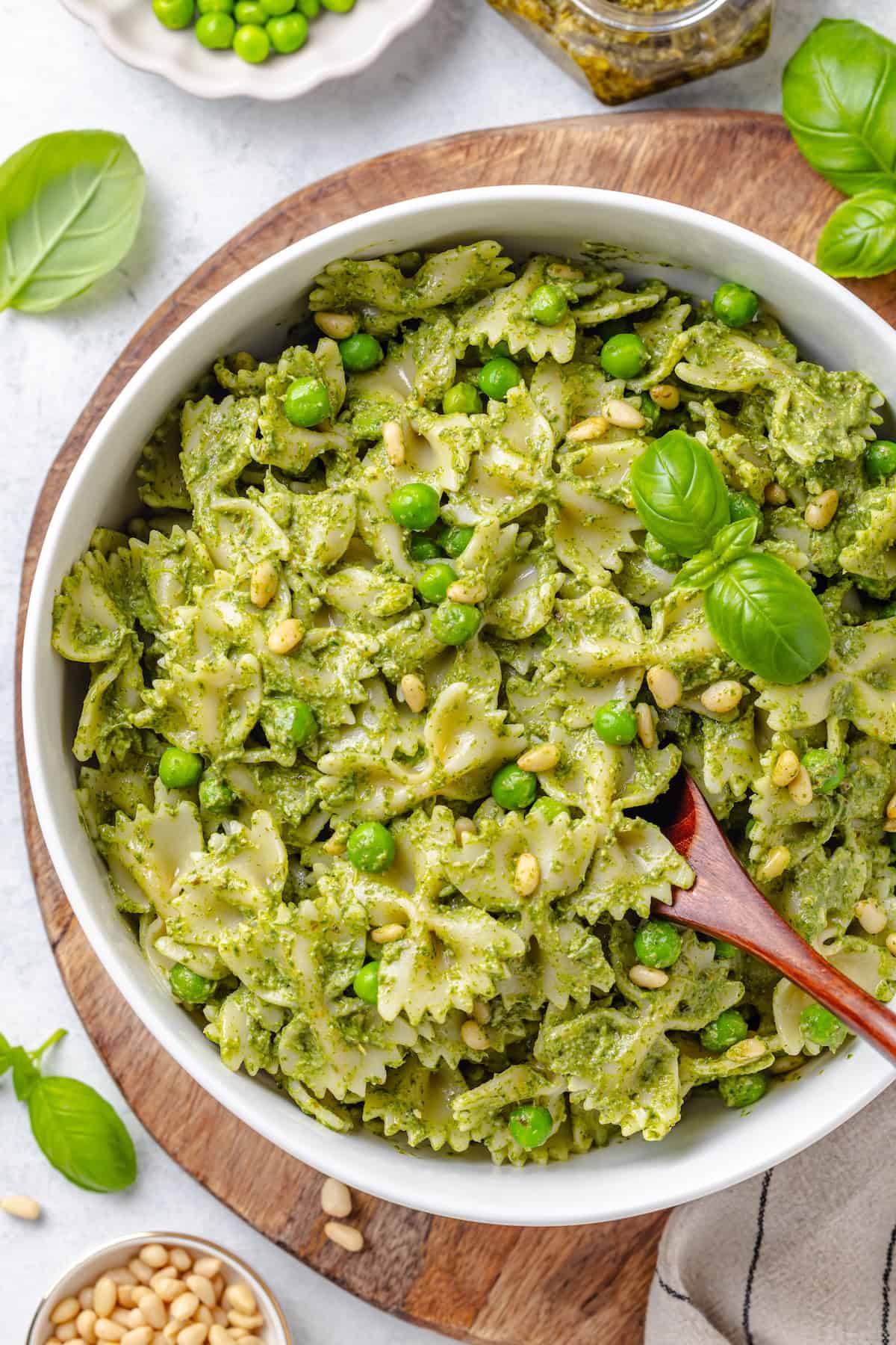 A pasta dish with pesto and peas in a large serving bowl with a wooden spoon resting in it.