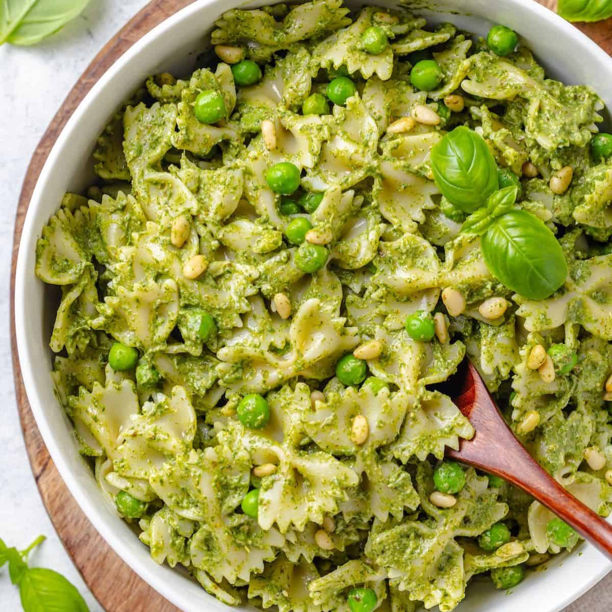 Close up of a pasta dish with pesto and peas in a large serving bowl with a wooden spoon resting in it.