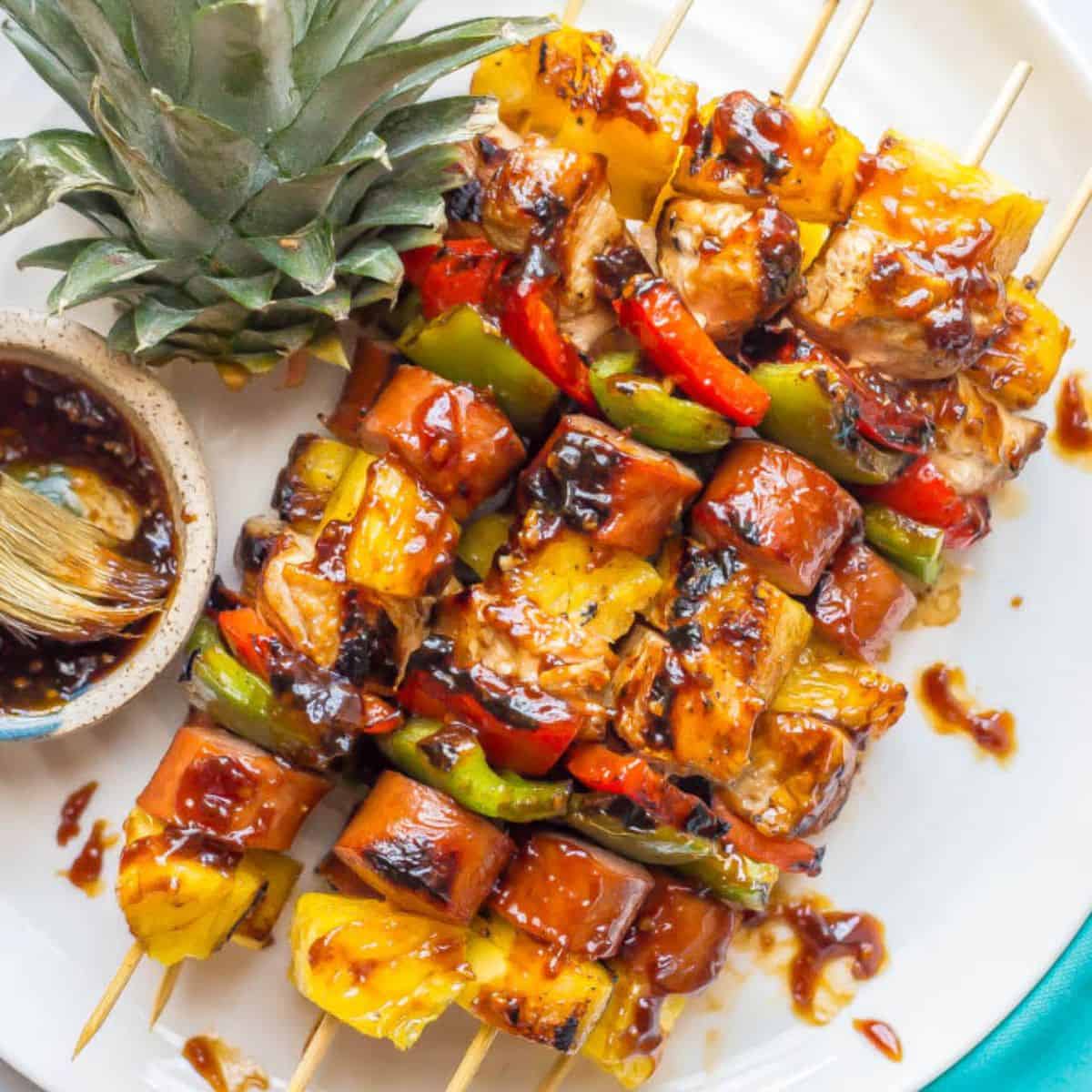 Close up of a serving plate with stacked kabobs with chicken, sausage, pepper and pineapple pieces brushed with teriyaki sauce.