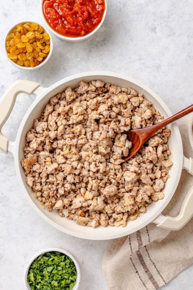 A deep, white skillet with a mixture of cooked, crumbled ground chicken and onions and spices.