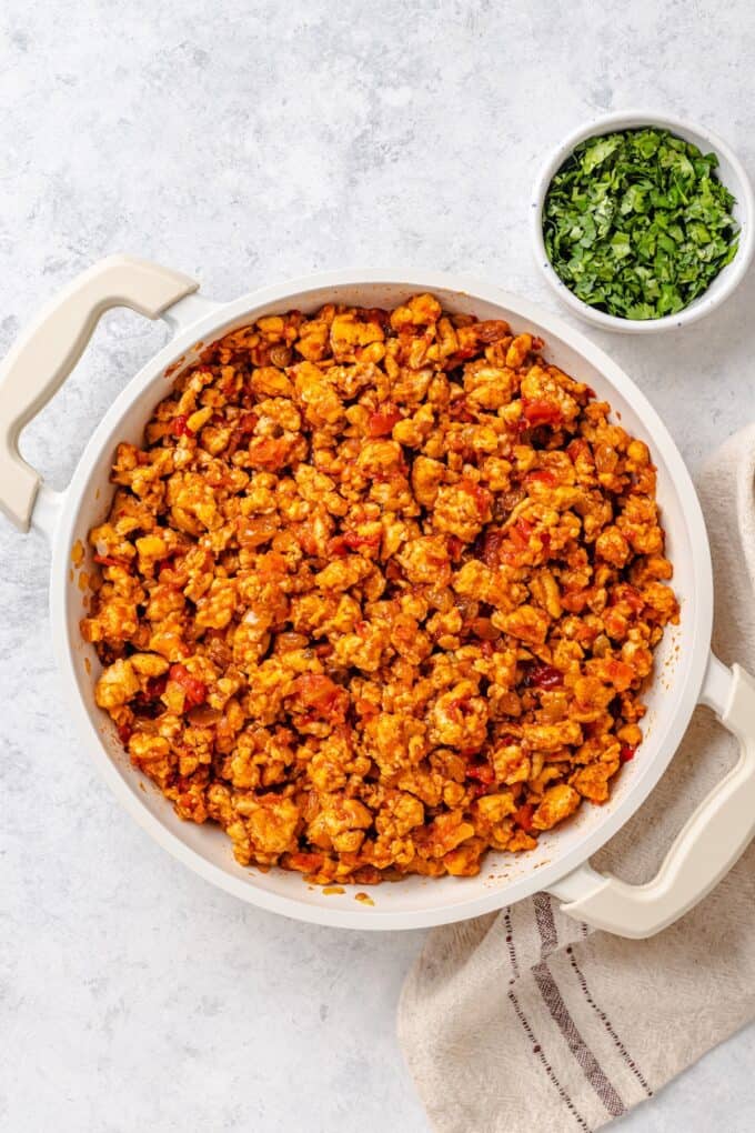 A spiced tomato and ground chicken mixture in a large deep, skillet with chopped fresh cilantro in a bowl to the side.