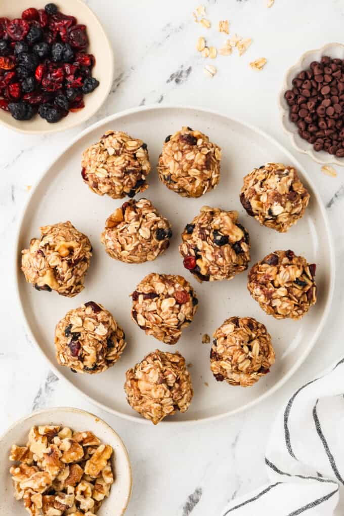 A white plate full of homemade granola bites with bowls of dried fruit and mini chocolate chips and walnuts nearby.