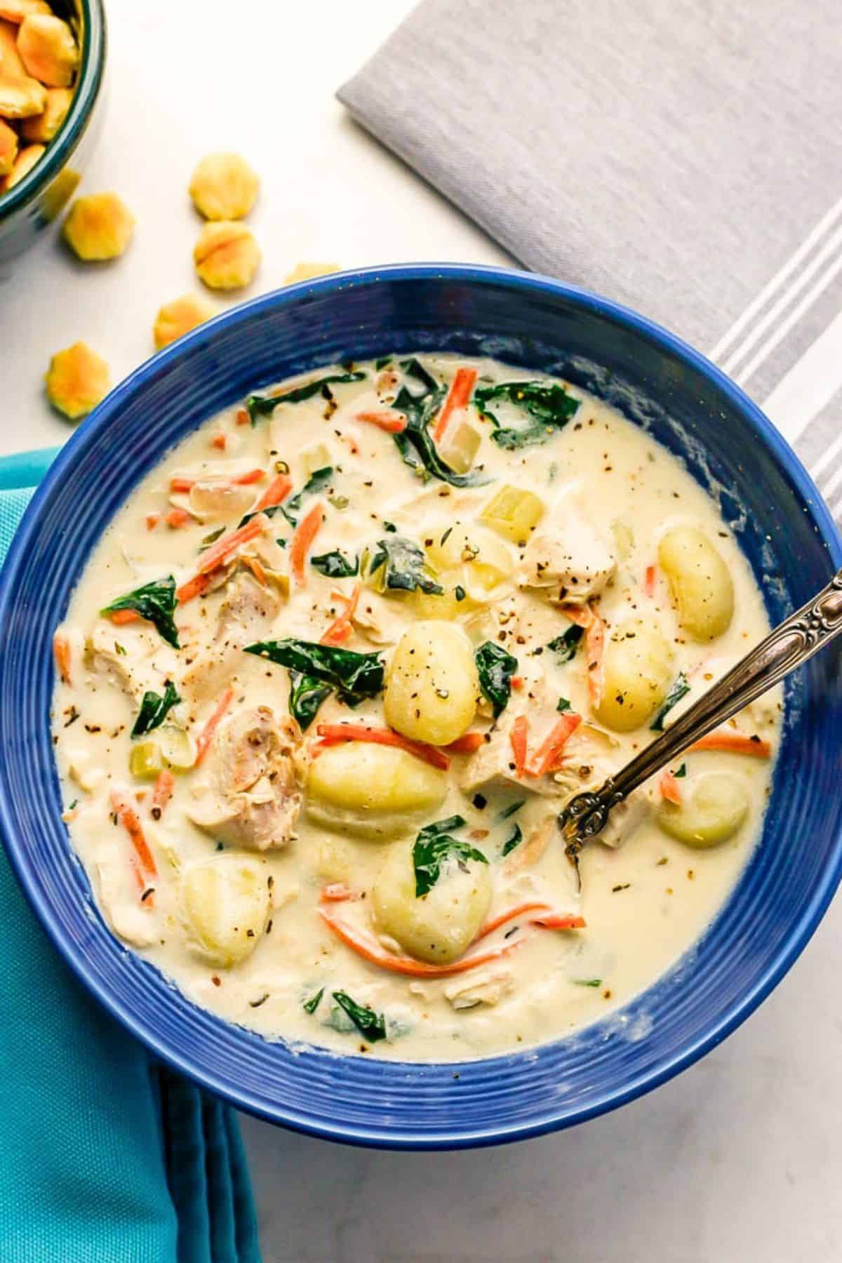 Chicken gnocchi soup is so creamy and comforting, easy to make and comes together in less than 30 minutes! This hearty soup is sure to warm you up. A blue bowl filled with a creamy chicken gnocchi soup with spinach and carrots and a spoon resting in it.