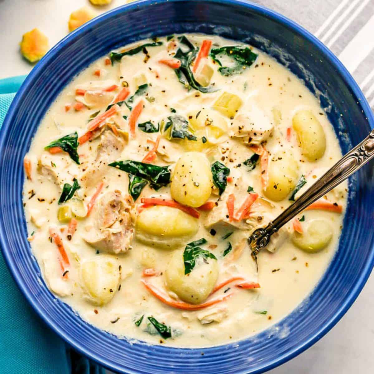Close up of a blue bowl filled with a creamy chicken gnocchi soup with spinach and carrots and a spoon resting in it.
