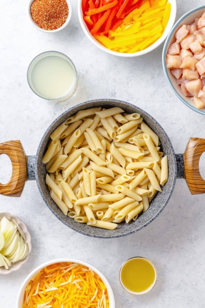 Cooked penne pasta in a grey bowl with bowls of other ingredients to the side.
