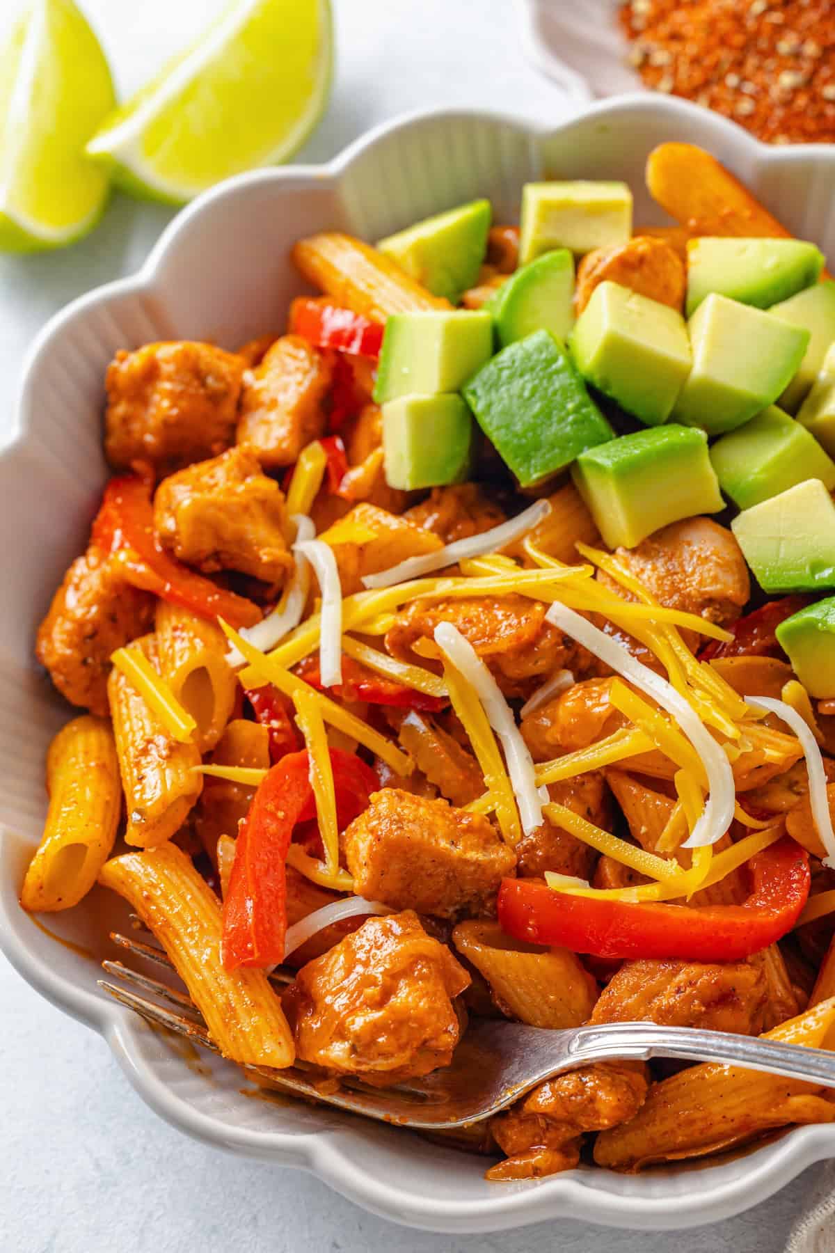 A fluted white bowl filled with a fajita chicken pasta mix topped with diced avocado and shredded cheese.