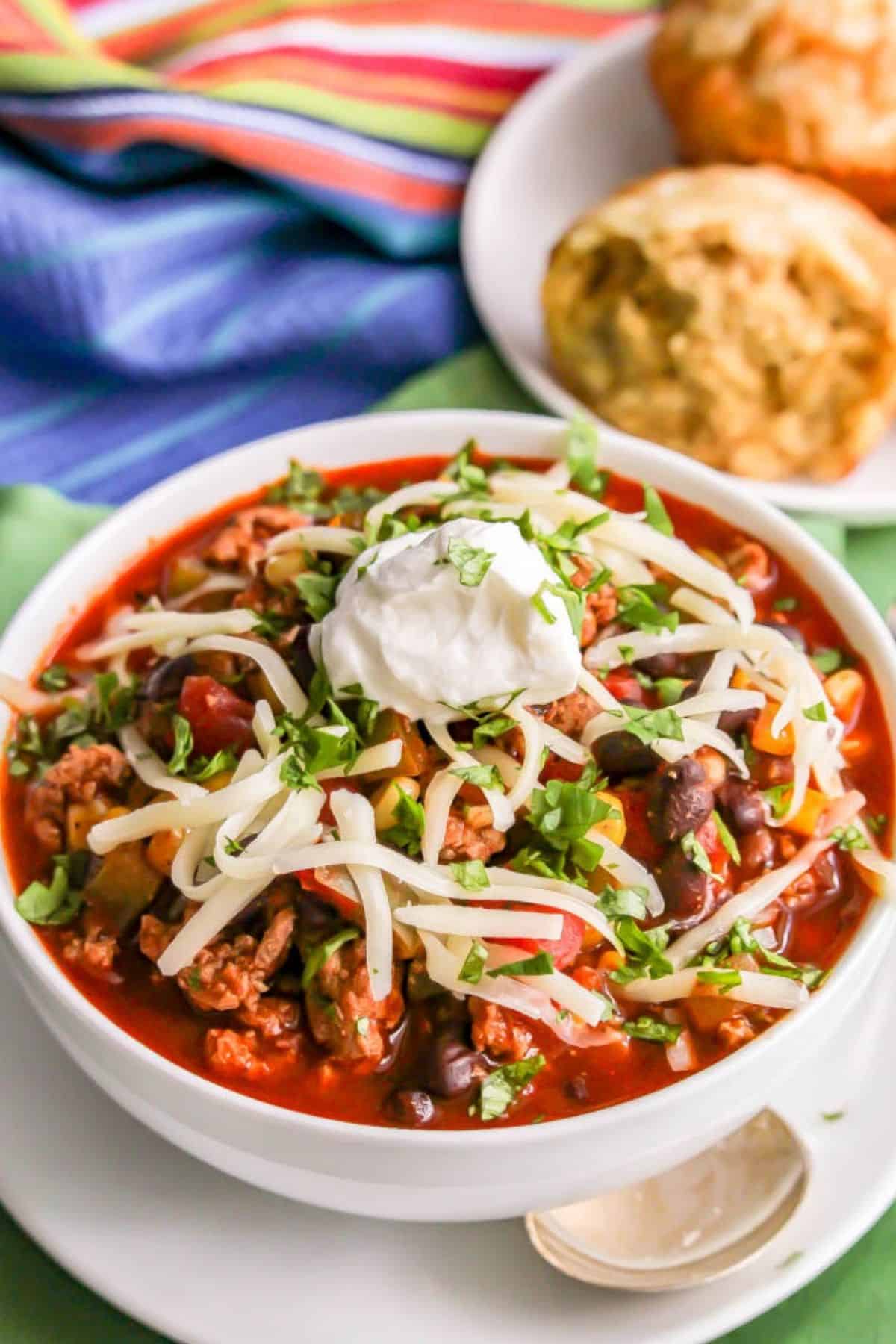 A bowl full of a southwest ground turkey chili with black beans and corn, plus sour cream and cheese on top, with a bowl of homemade drop biscuits in the background.