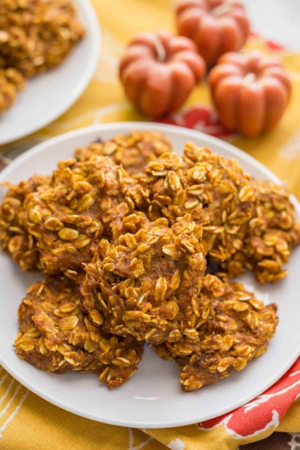 A white plate with a stack of pumpkin banana oatmeal cookies with some pumpkin shaped candles in the background.