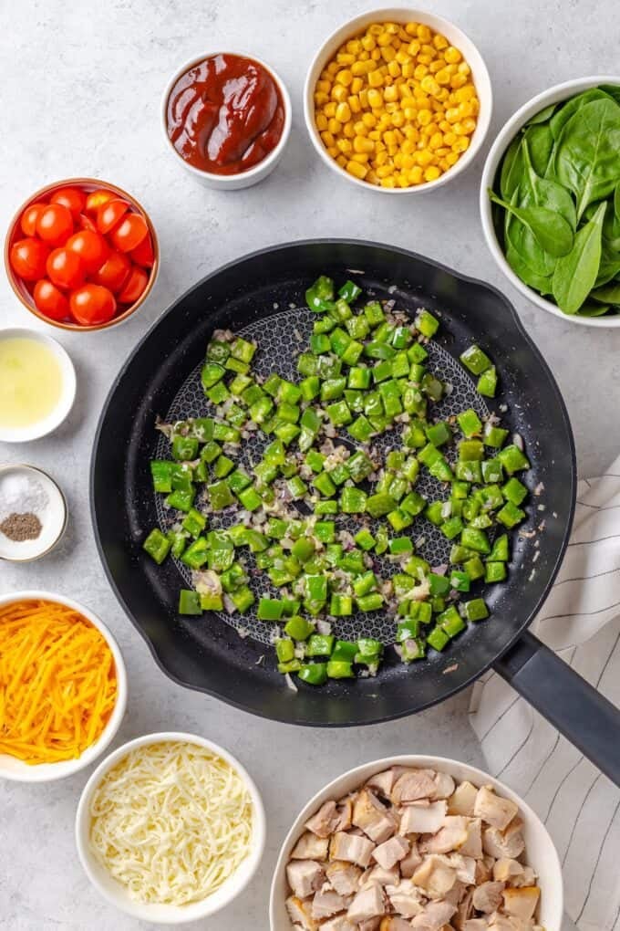 Red onion and green pepper being sautéed in a large cast iron skillet with other ingredients in bowls around the perimeter.