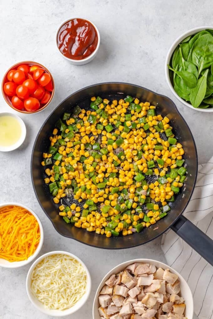 Red onion and green pepper and corn being sautéed in a large cast iron skillet with other ingredients in bowls around the perimeter.