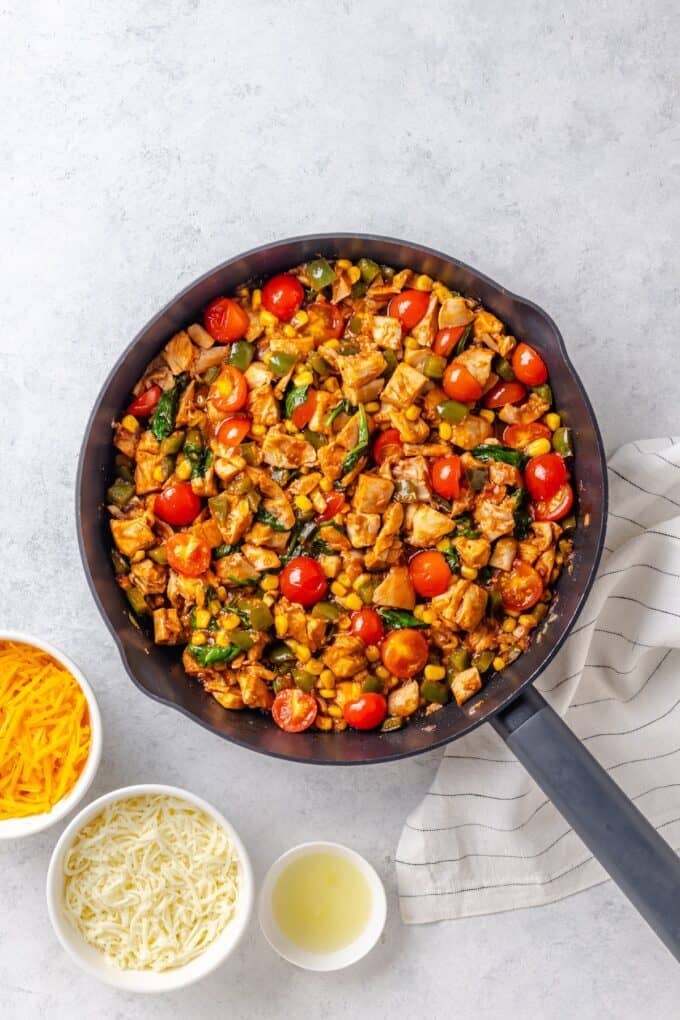 A large cast iron skillet with a mixture of BBQ chicken, corn, tomatoes and wilted spinach.