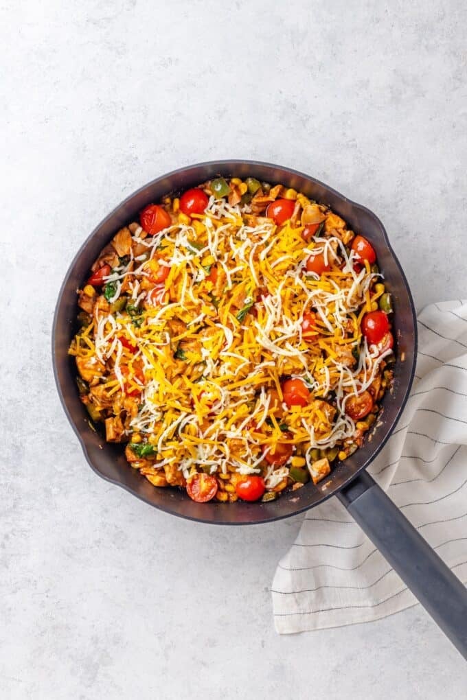 A large cast iron skillet with a mixture of BBQ chicken, corn, tomatoes and wilted spinach topped with a mix of shredded mozzarella and cheddar cheese.