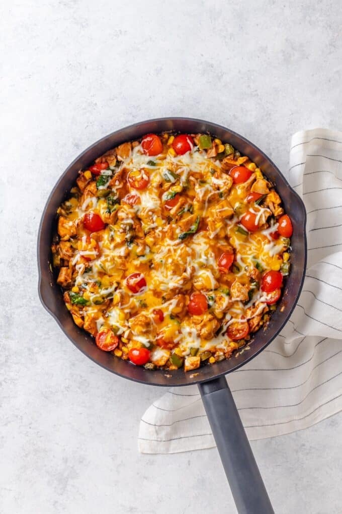 A large cast iron skillet with a mixture of BBQ chicken, corn, tomatoes and wilted spinach with melted cheese on top.