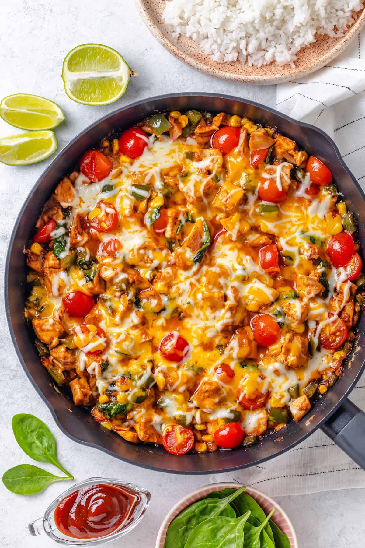 A BBQ chicken skillet with spinach and tomatoes and melted cheese on top with a bowl of rice and some lime wedges to the side.