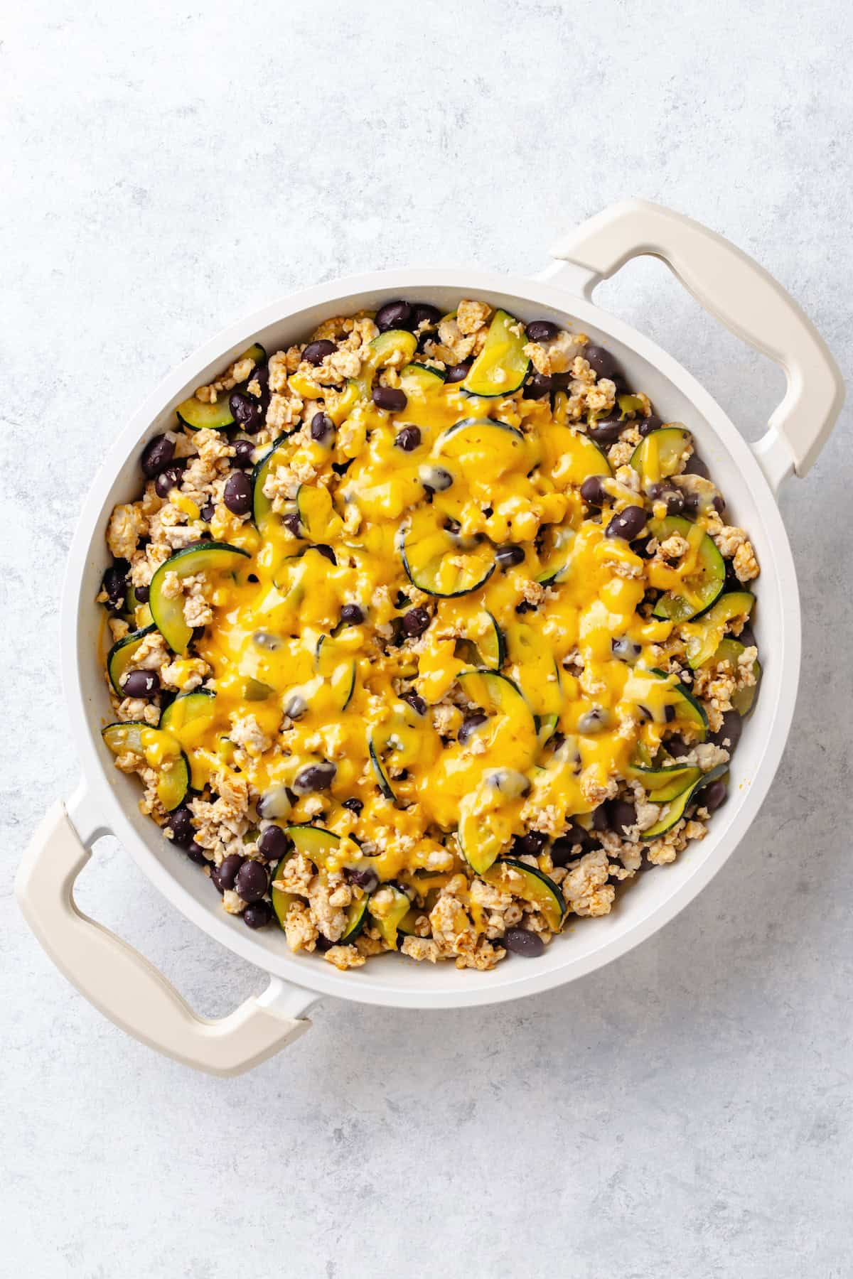 Melted cheese on top of a mixture of ground turkey and zucchini with black beans in a large white skillet.