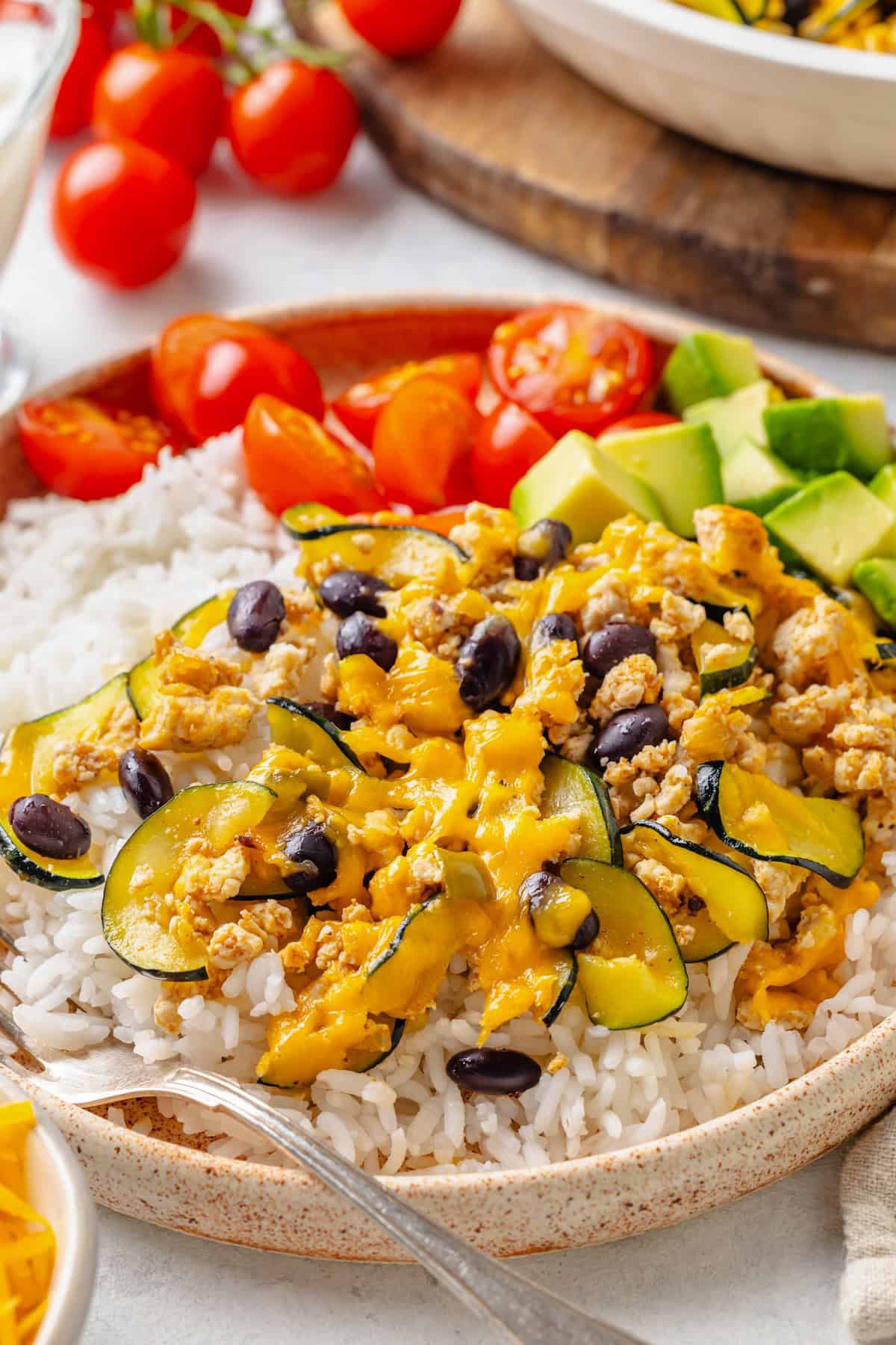 A dinner plate with white rice topped with a ground turkey mixture with zucchini and black beans and cheese on top, with avocado and tomatoes to the side.