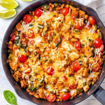 Close up of A BBQ chicken skillet with spinach and tomatoes and melted cheese on top with some lime wedges to the side.