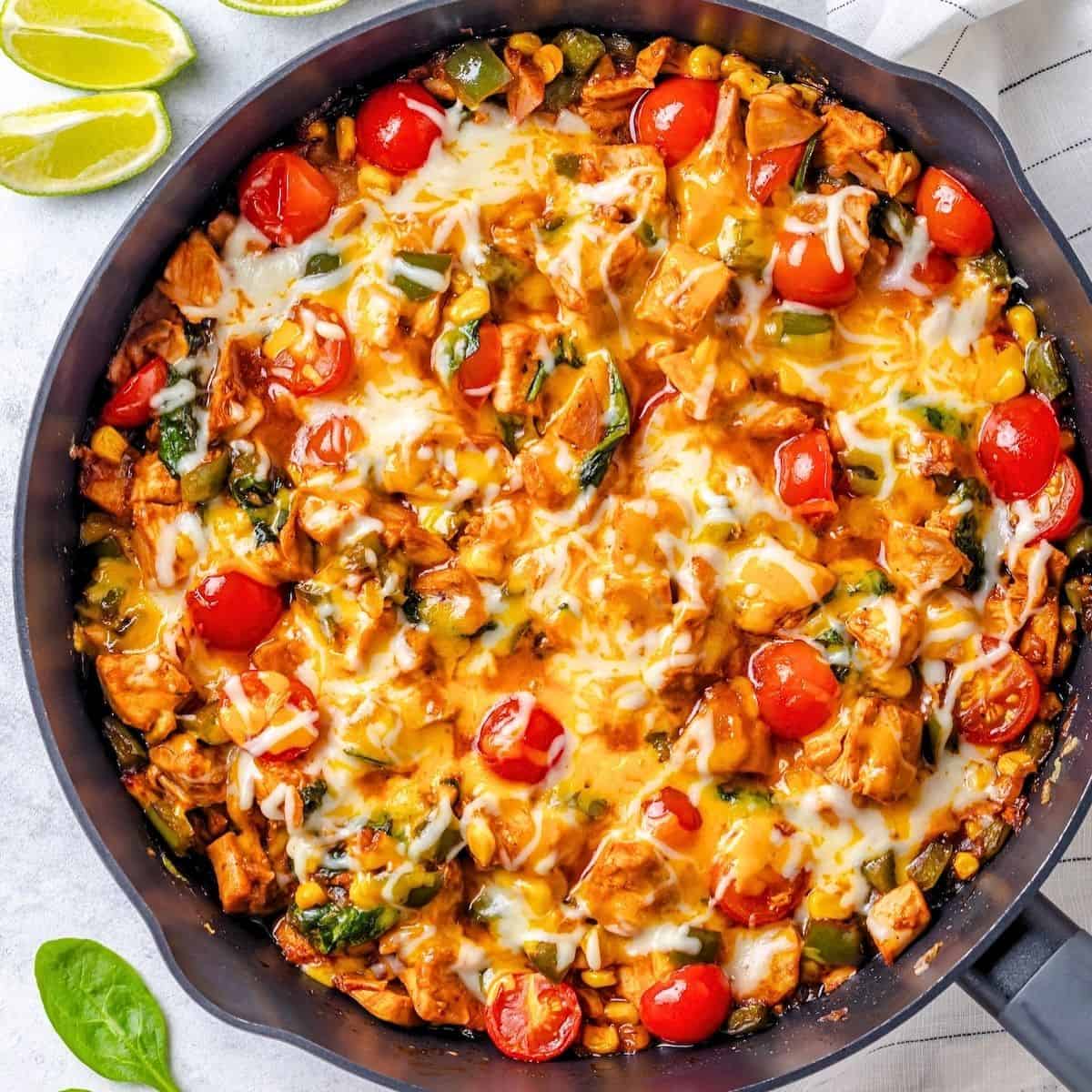 Close up of A BBQ chicken skillet with spinach and tomatoes and melted cheese on top with some lime wedges to the side.