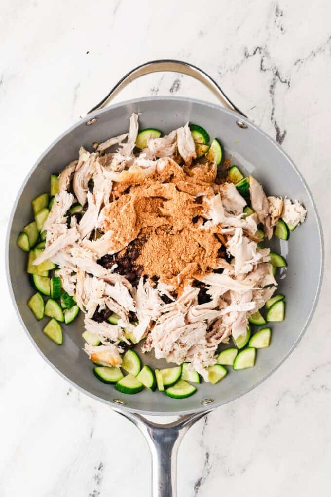 A large skillet with zucchini, rotisserie chicken, black beans and some seasoning on top before being stirred together.