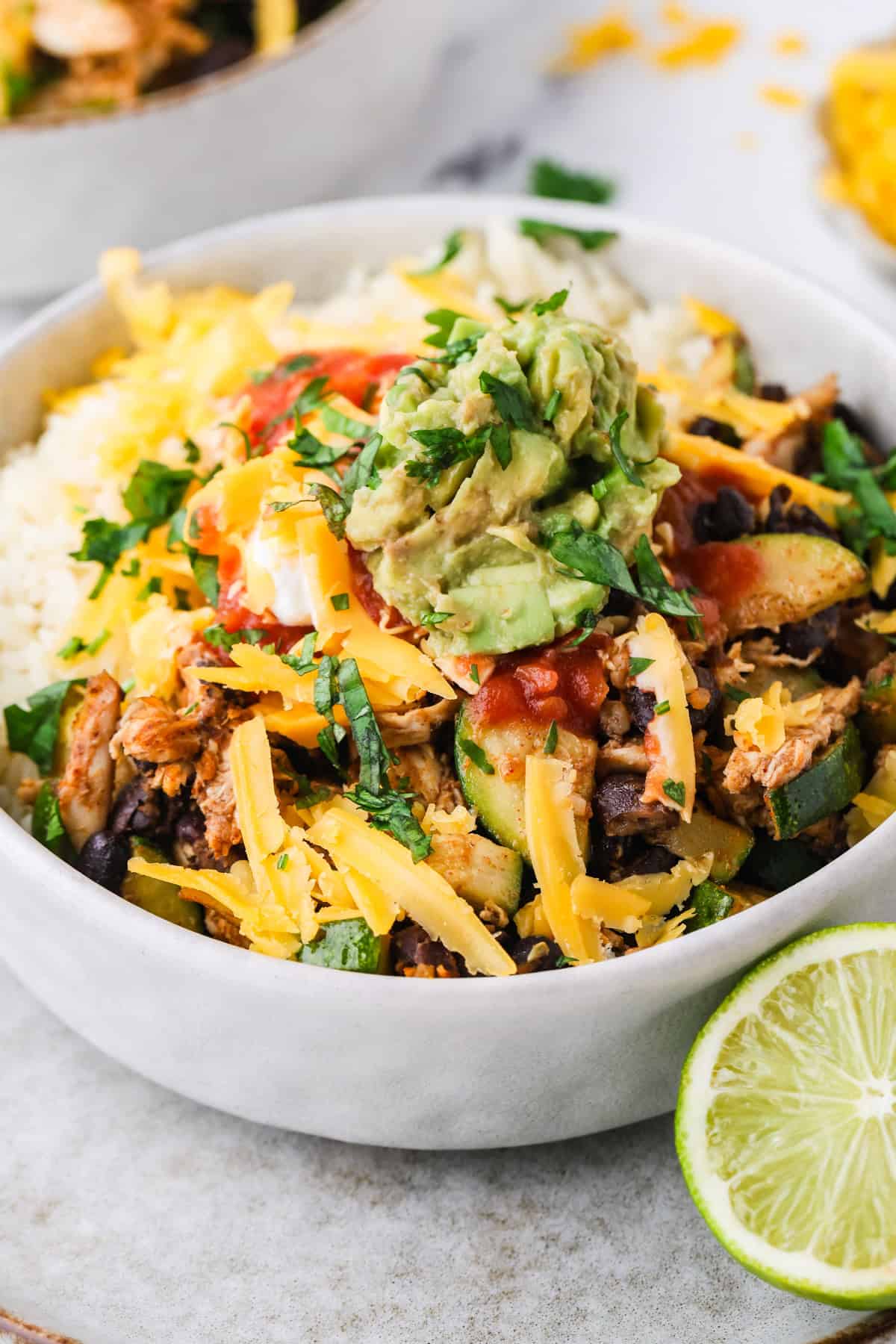 Close up of a rice bowl with a chicken, black bean and zucchini mixture and cheese, salsa and guac on top.