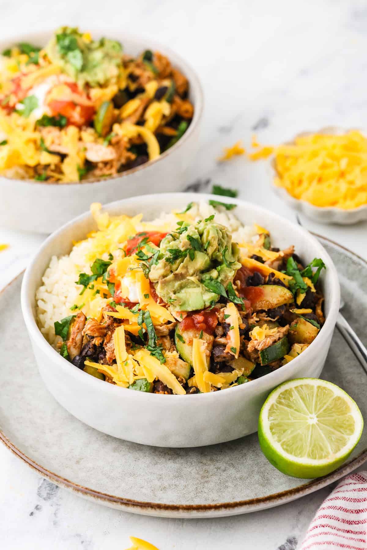Two white bowls with rice alongside a chicken, black bean and zucchini mixture and cheese, salsa and guac on top.