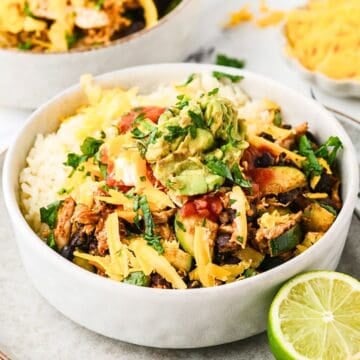 Close up of a white bowl with rice alongside a chicken, black bean and zucchini mixture and cheese, salsa and guac on top.