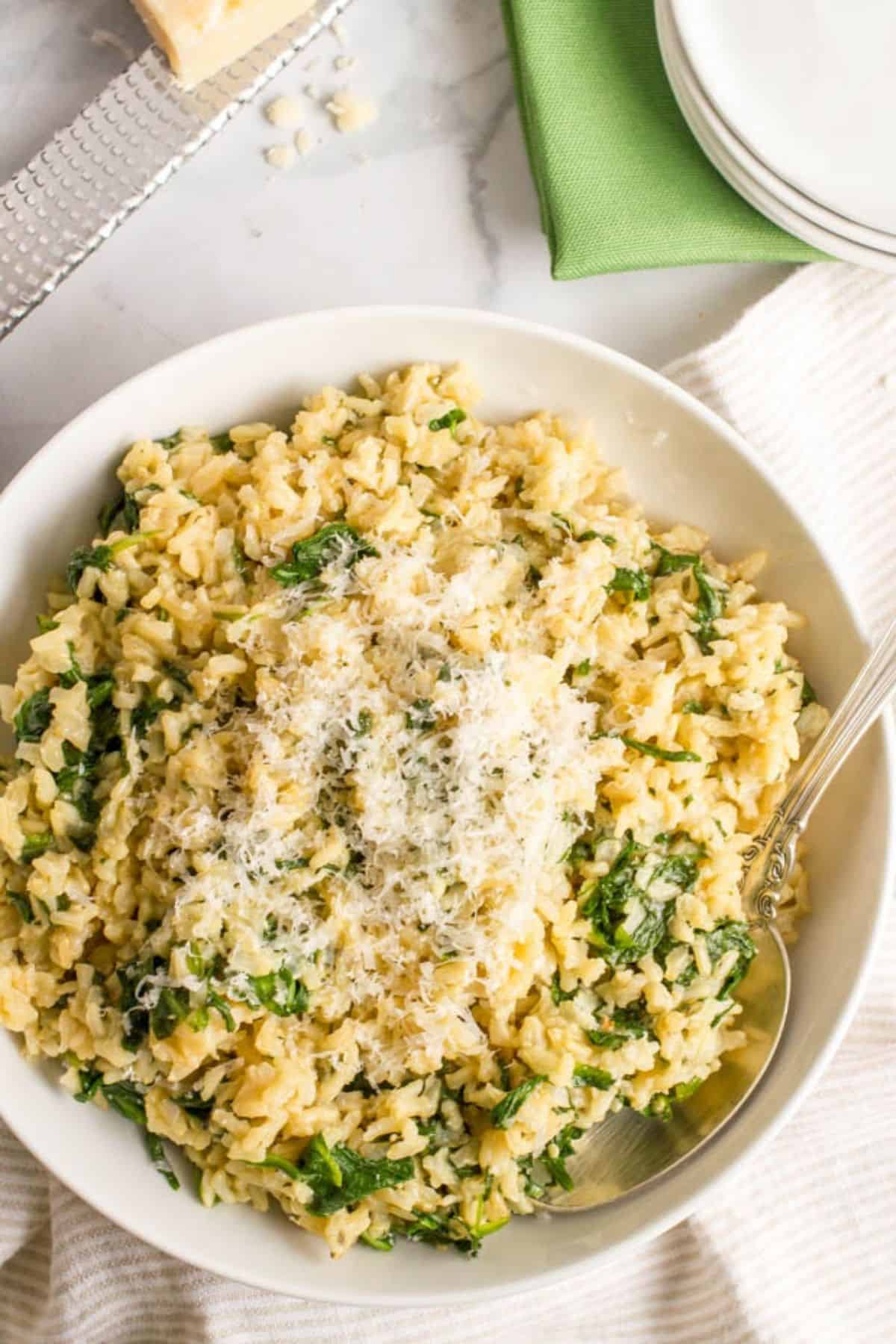 Brown rice with spinach and Parmesan cheese is an easy, healthy, one-pot side dish with just a few simple ingredients! #brownrice #wholegrains #easysidedish #glutenfreerecipe A white serving bowl full of brown rice mixed with wilted spinach and topped with Parmesan cheese with a spoon resting in it and plates to the side.