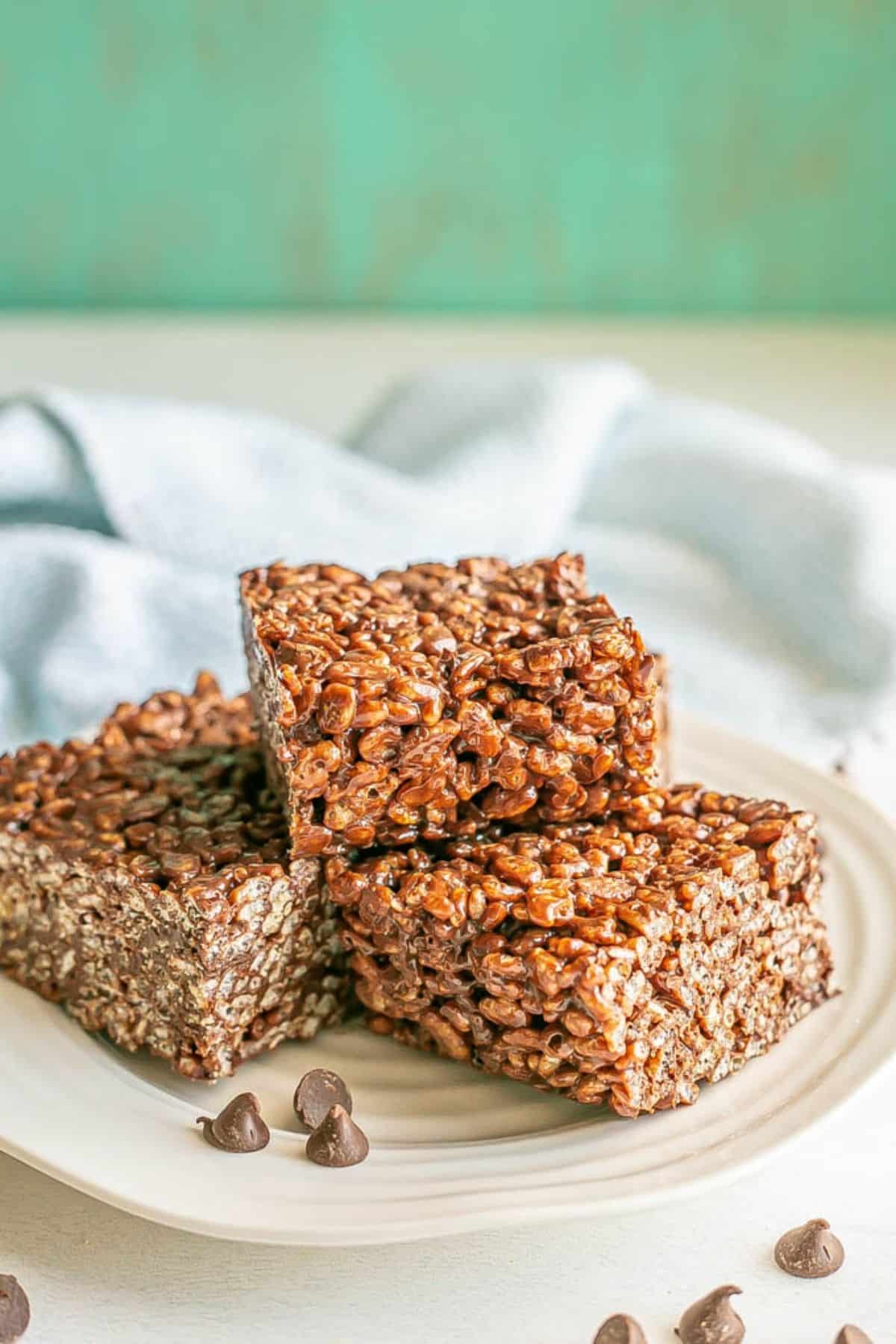 A stack of chocolate rice krispies squares on a white plate with chocolate chips sprinkled nearby.
