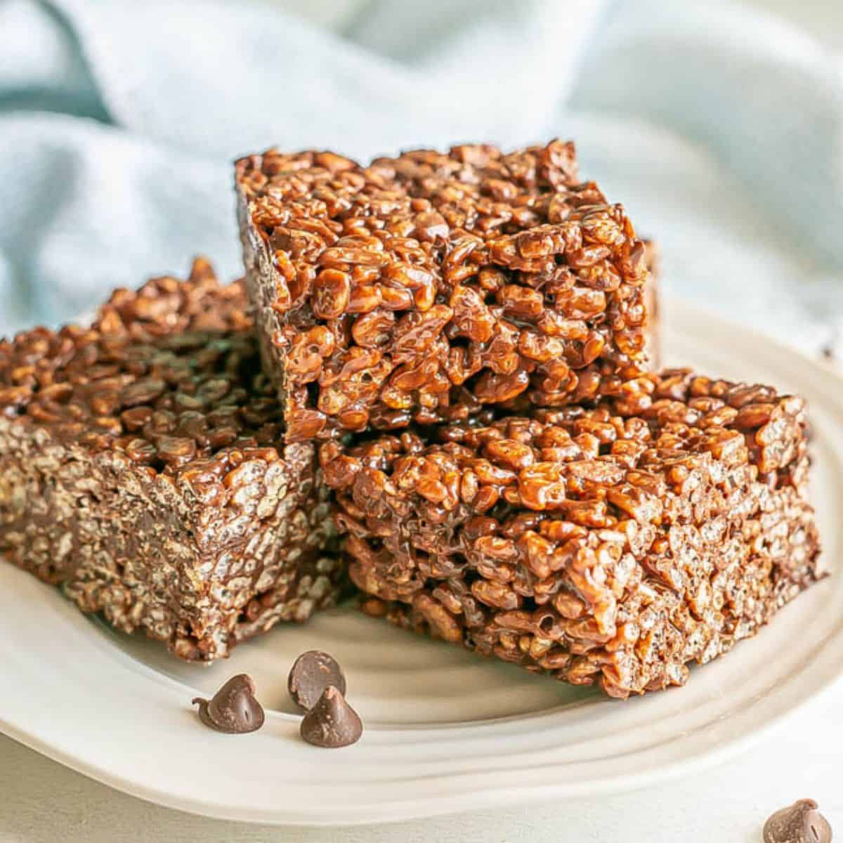 Close up of a stack of chocolate rice krispies squares on a white plate with chocolate chips sprinkled nearby.