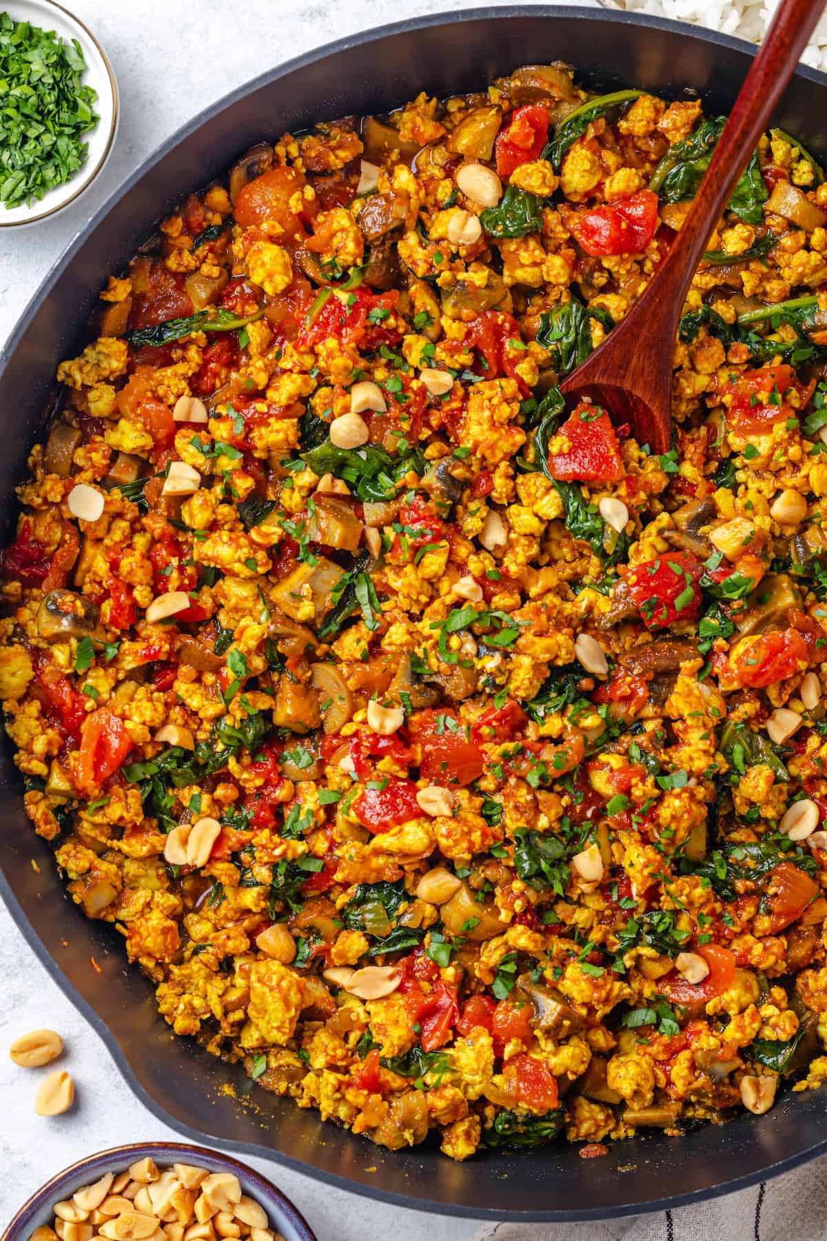 A wooden spoon resting in a large dark skillet with a curried ground chicken mixture with spinach and tomatoes.