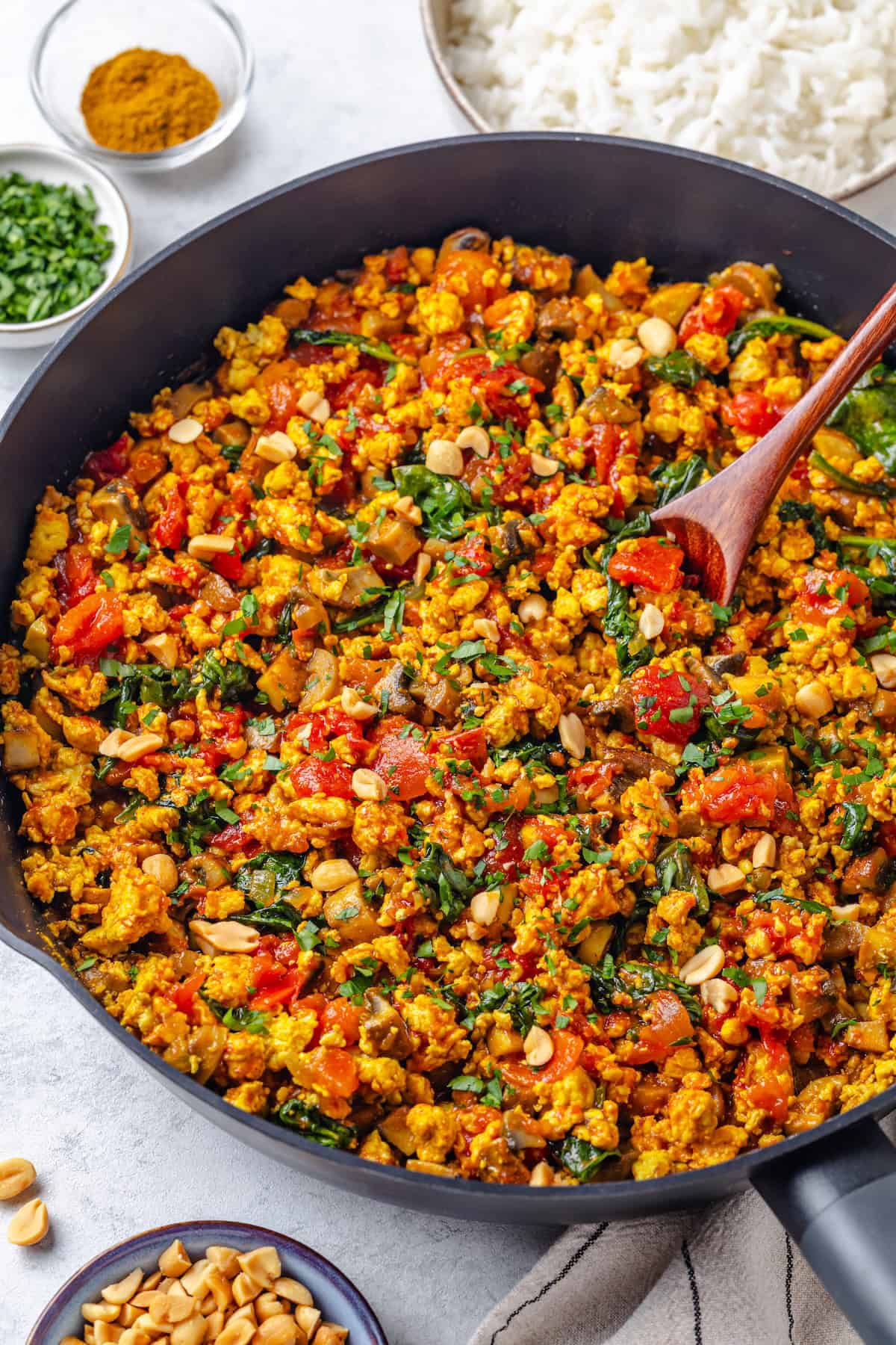 A wooden spoon resting in a large skillet filled with a curried ground chicken with mushrooms, spinach and tomatoes.