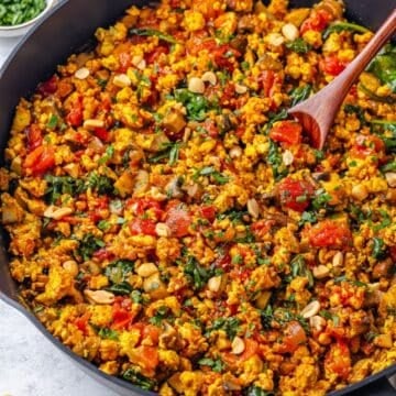 Close up of a wooden spoon resting in a large skillet filled with a curried ground chicken with mushrooms, spinach and tomatoes.
