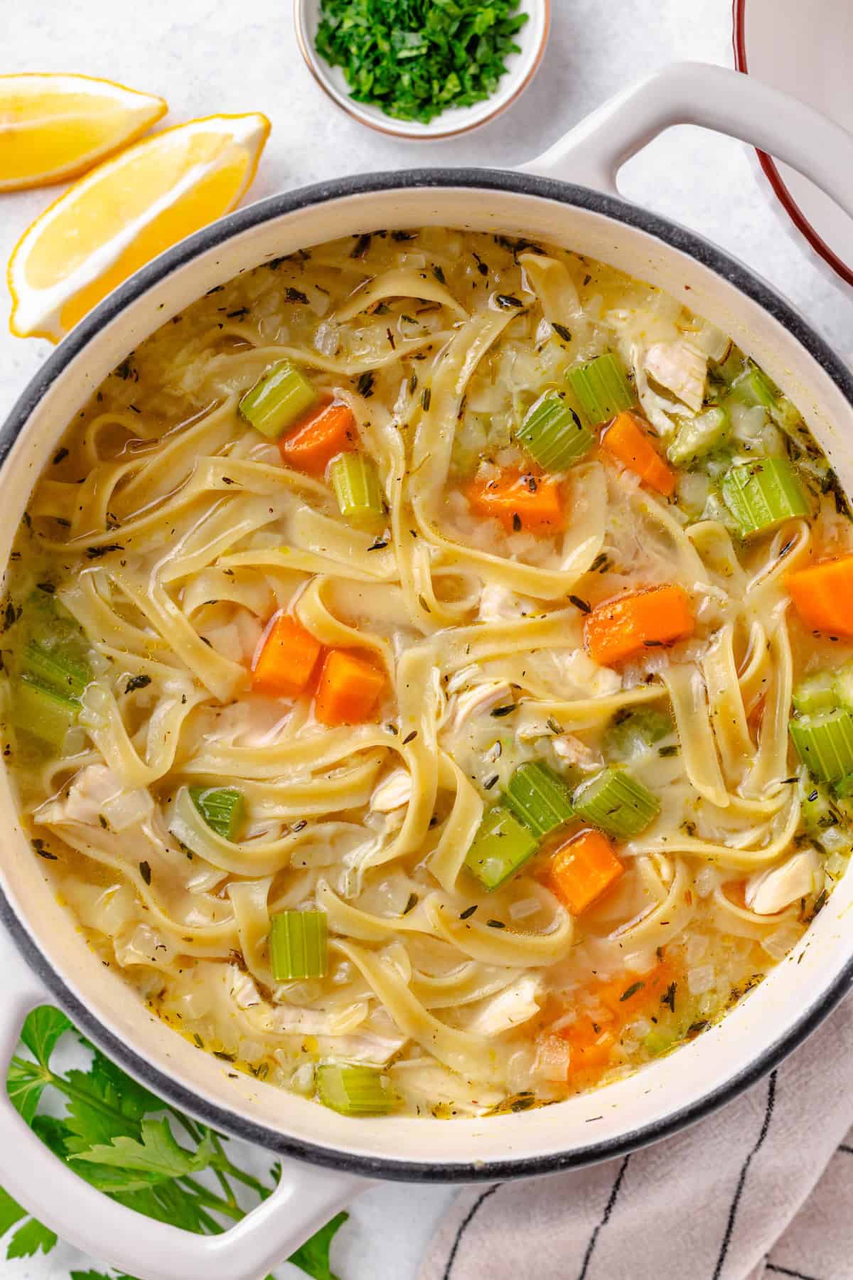 A dutch oven filled with a simple homemade chicken noodle soup with lemon wedges and fresh herbs to the side.