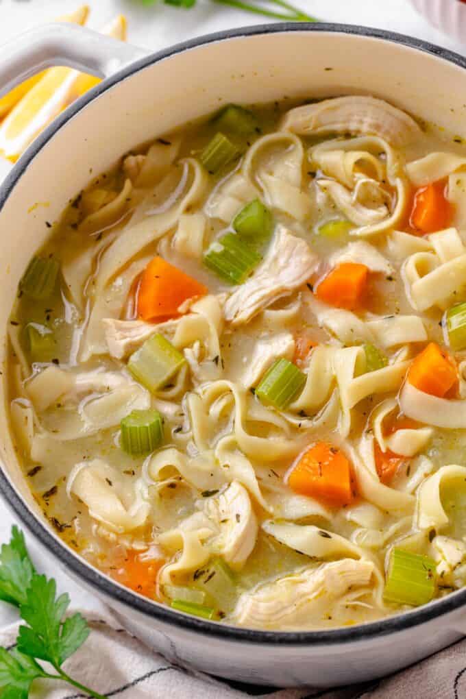 Close up of a dutch oven filled with a simple homemade chicken noodle soup with lemon wedges and fresh herbs to the side.