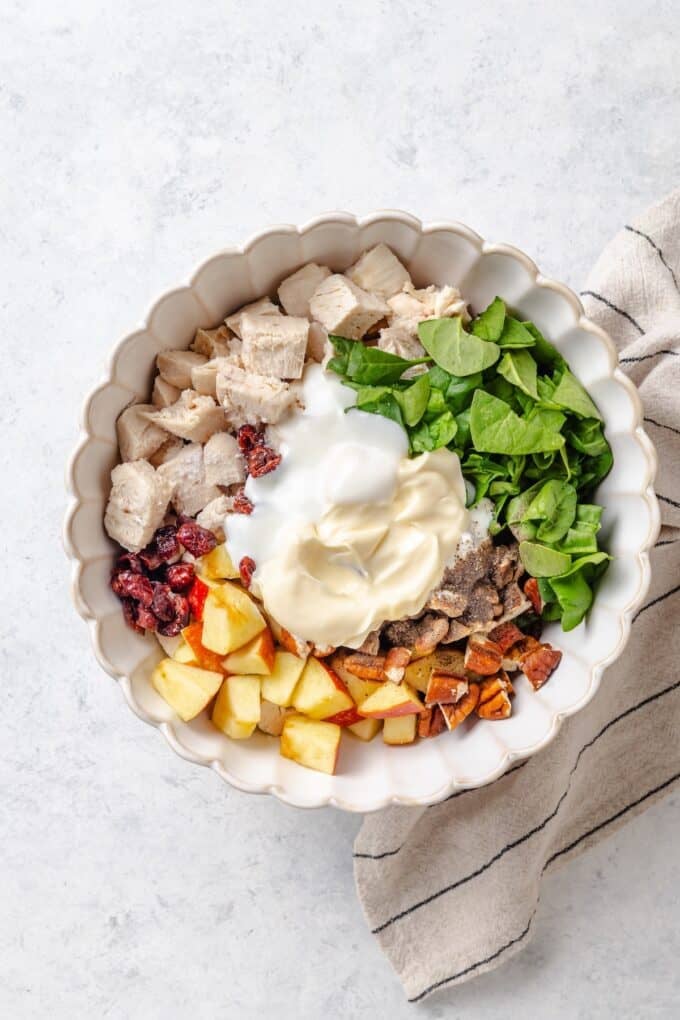 A large fluted bowl with separate piles of cooked chicken, chopped spinach, pecans, apple chunks and dried cranberries with a dressing mixture of mayonnaise and Greek yogurt in the middle before being stirred together.