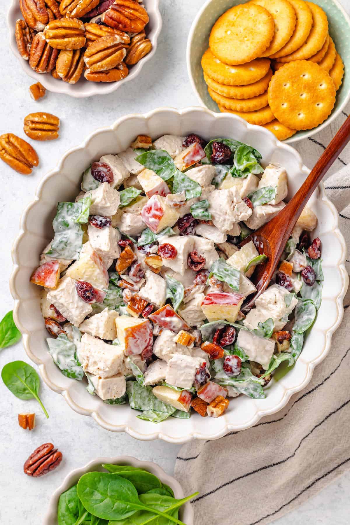 A spoon resting in a large fluted serving bowl filled with a harvest chicken salad with apples and pecans with bowls of pecans and crackers nearby.