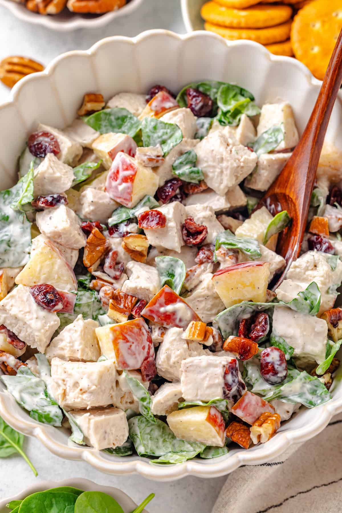Harvest chicken salad with pecans, apples and dried cranberries is a delicious, crunchy, fresh fall twist on the classic! It's great served as a sandwich, wrap or with crackers or greens. #chickensalad #lunchrecipes #fallrecipes #lunchtime #chickenrecipes Close up of a chicken salad mixture with apples, pecans and spinach in a large fluted bowl with a wooden spoon resting in it.