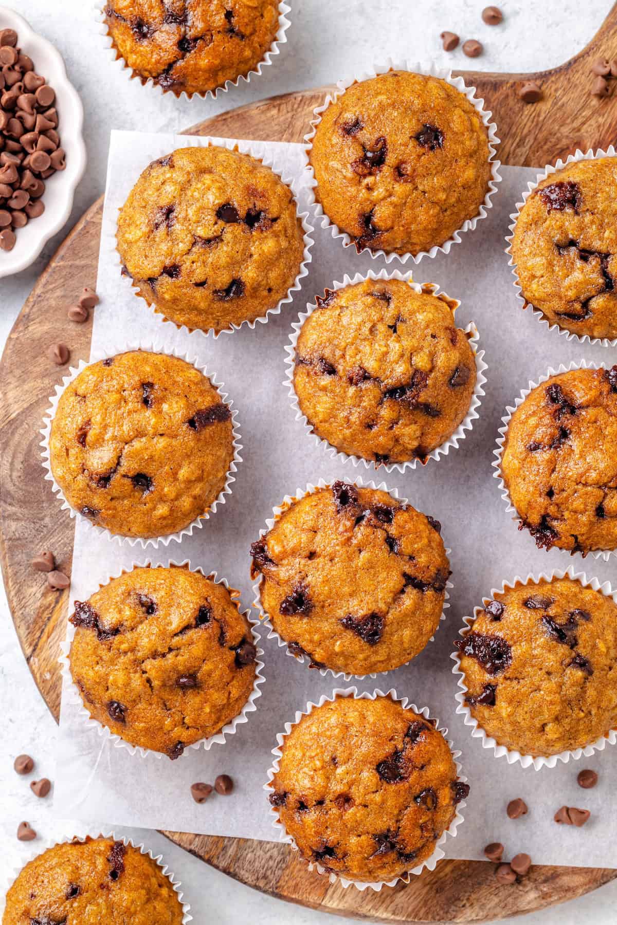 A cutting board with a piece of parchment paper and an array of healthy chocolate chip muffins set out.