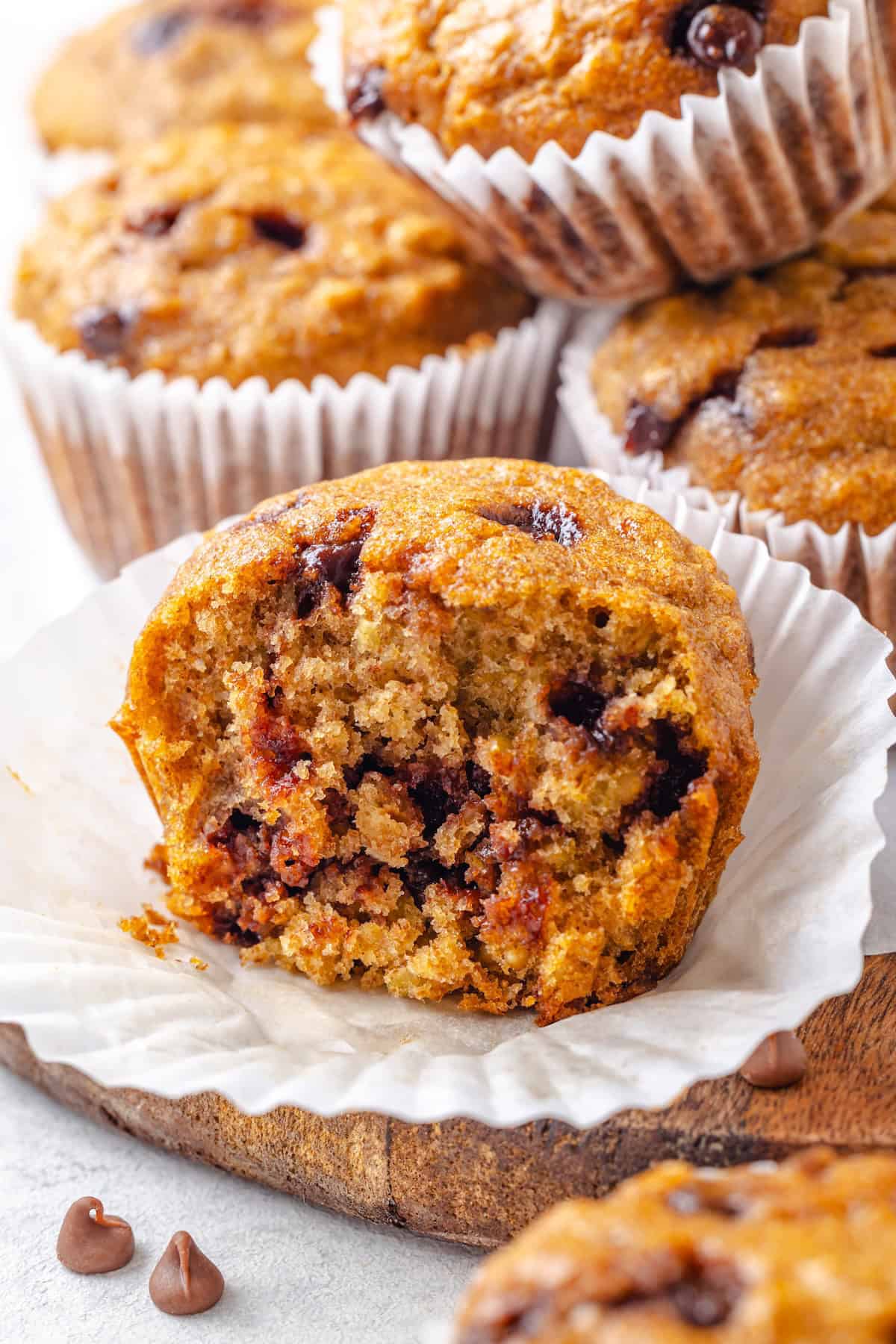 A chocolate chip muffin with a bite taken out of it resting on a muffin liner on a wood tray.