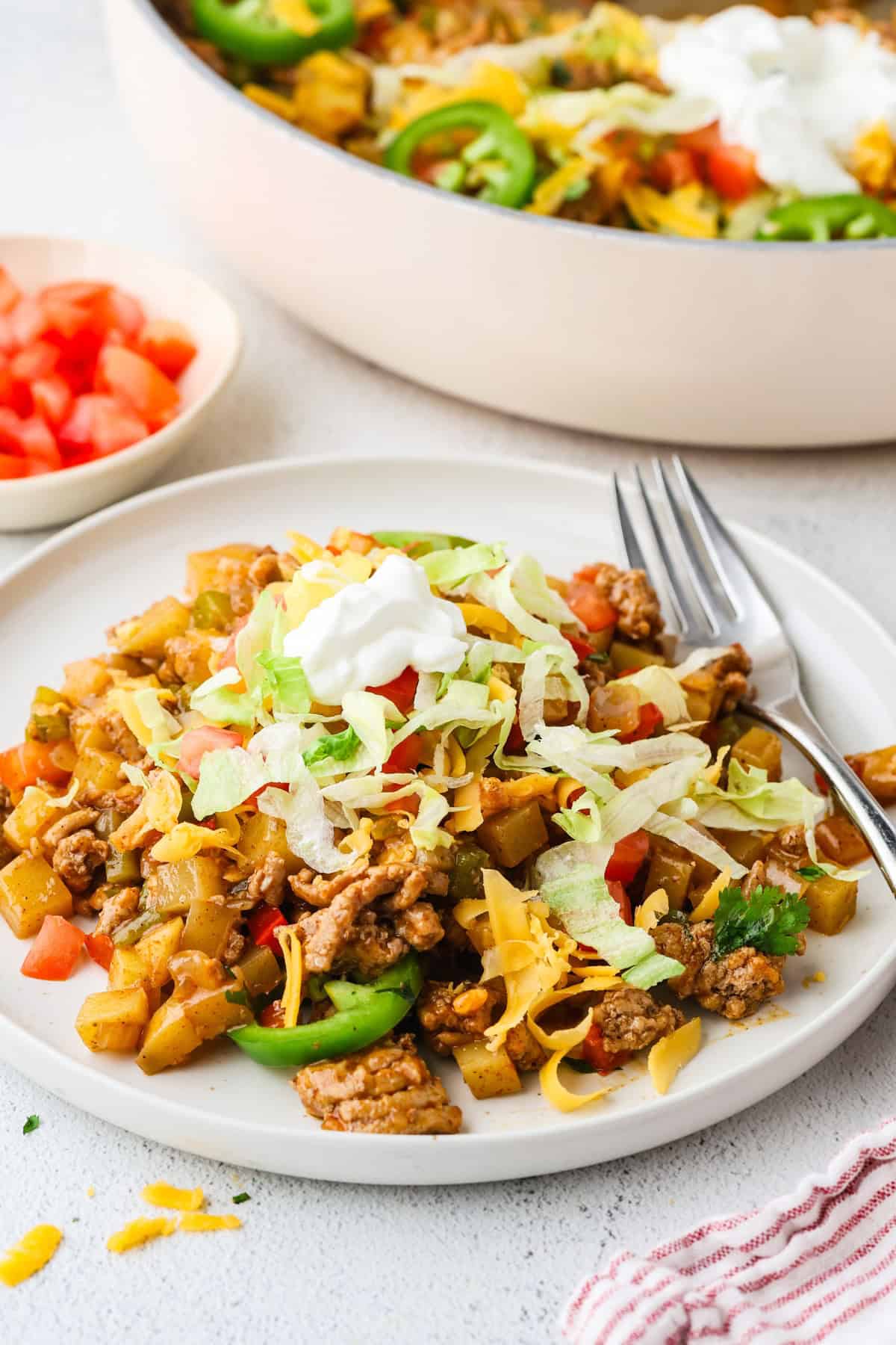 A white dinner plate with a serving of taco turkey and potatoes with toppings of shredded lettuce, cheese and sour cream on top with a skillet in the background.