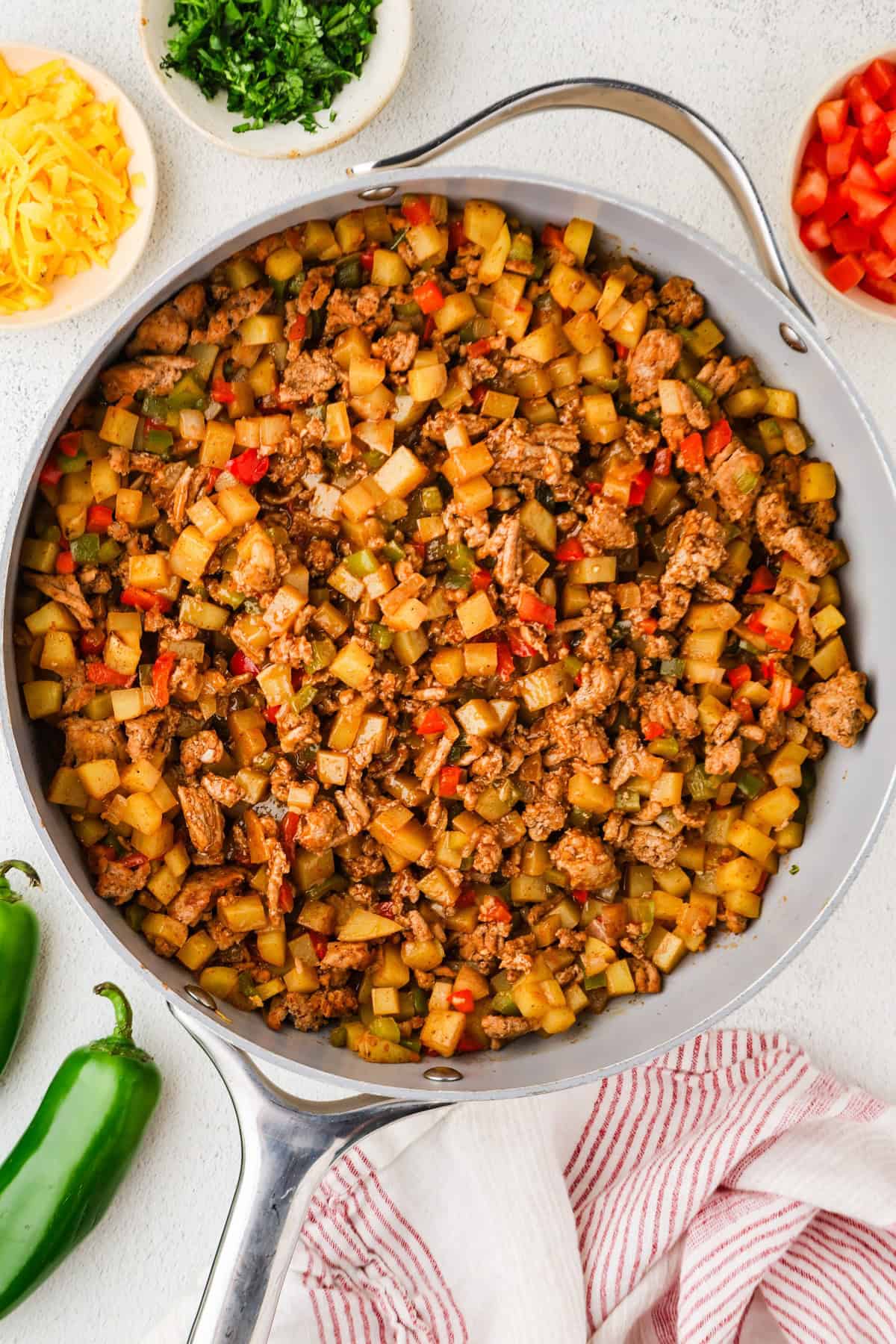 A large skillet full of cooked ground turkey, potatoes and peppers with taco seasoning with bowls of toppings to the side.
