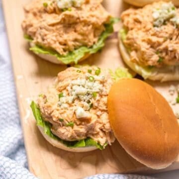 Close up of a wooden board loaded with sliders piled with a healthier slow cooker buffalo chicken mixture topped with blue cheese crumbles and sliced green onions.