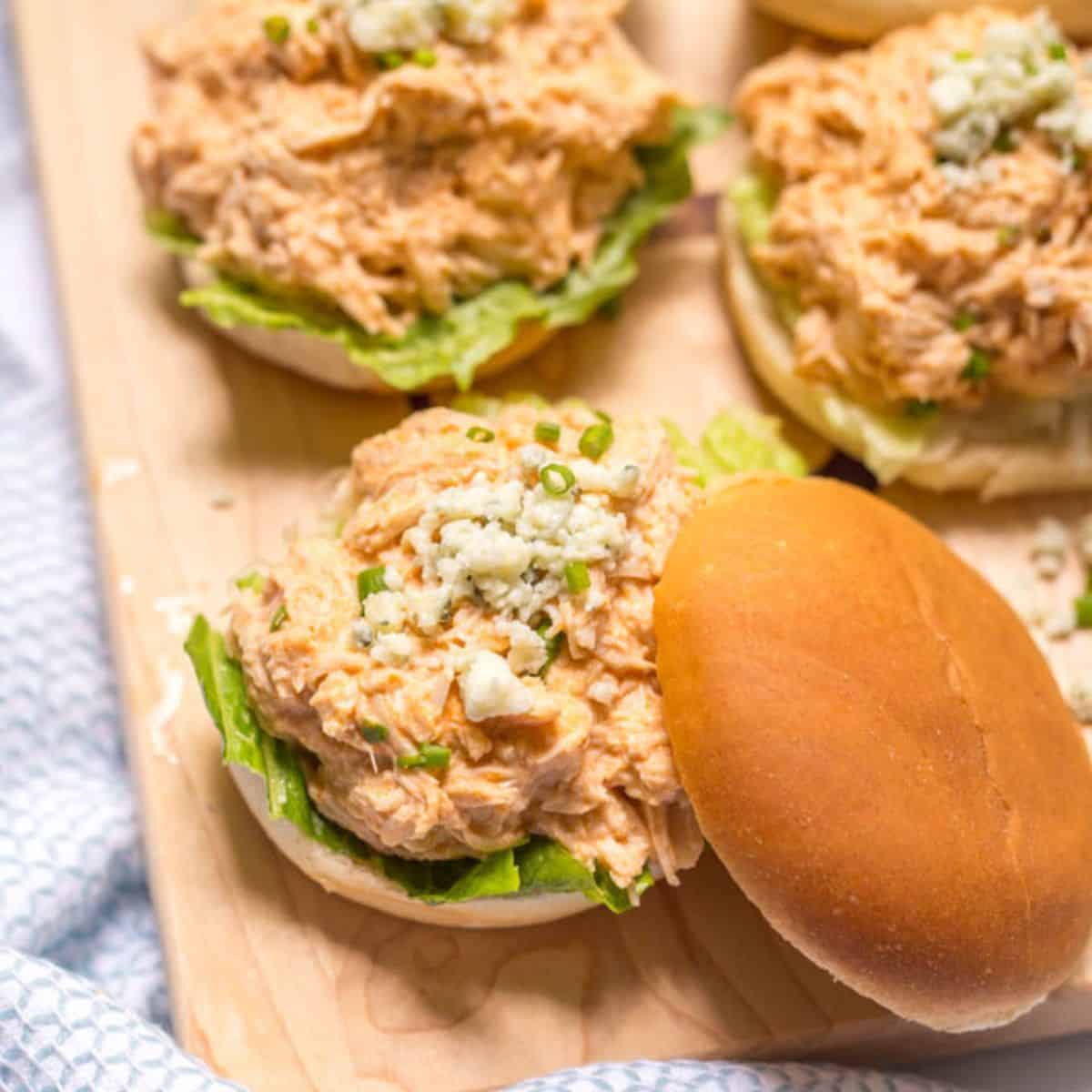 Close up of a wooden board loaded with sliders piled with a healthier slow cooker buffalo chicken mixture topped with blue cheese crumbles and sliced green onions.