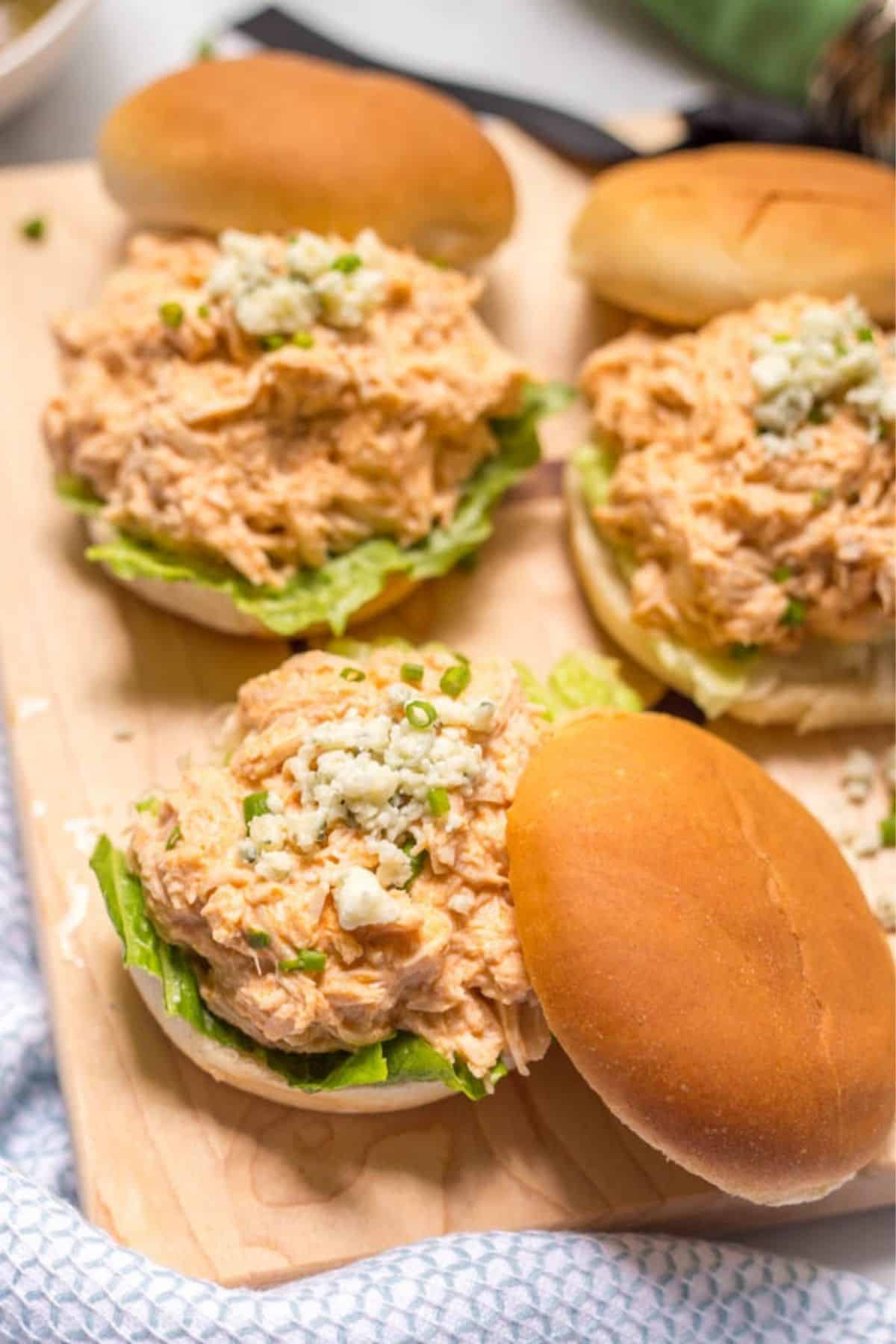 Healthy slow cooker buffalo chicken is lightened up (no butter!), takes just minutes to prep and can be used for sandwiches, sliders, wraps, salads or nachos! | www.familyfoodonthetable.com A wooden board loaded with sliders piled with a healthier slow cooker buffalo chicken mixture topped with blue cheese crumbles and sliced green onions.