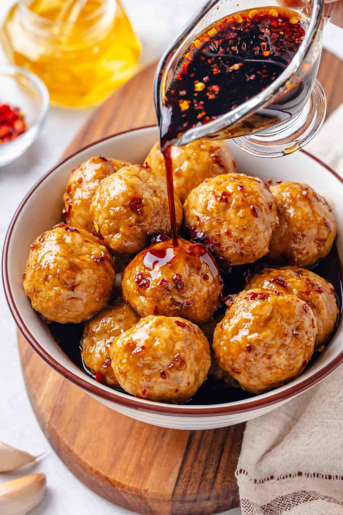 Honey garlic sauce being poured over meatballs in a white bowl with a red rim.