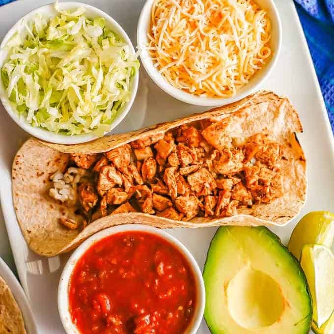 A rectangular white plate with a soft shell taco with rice and leftover turkey with bowls of toppings on the side